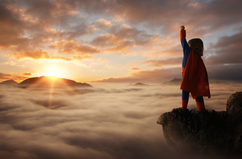 Kind im Superman Kostüm auf einem Berg über den Wolken mit Sonnenaufgang